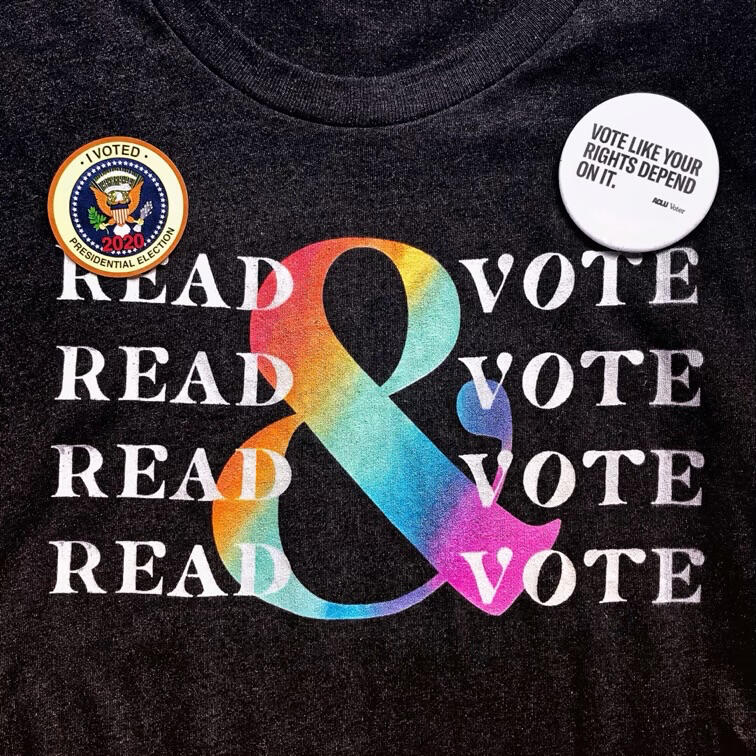 A dark grey shirt with the words “Read and Vote” printed four times in bold letters. An “I Voted” sticker and ACLU pin are attached. The pin reads, “Vote like your rights depends on it.”
