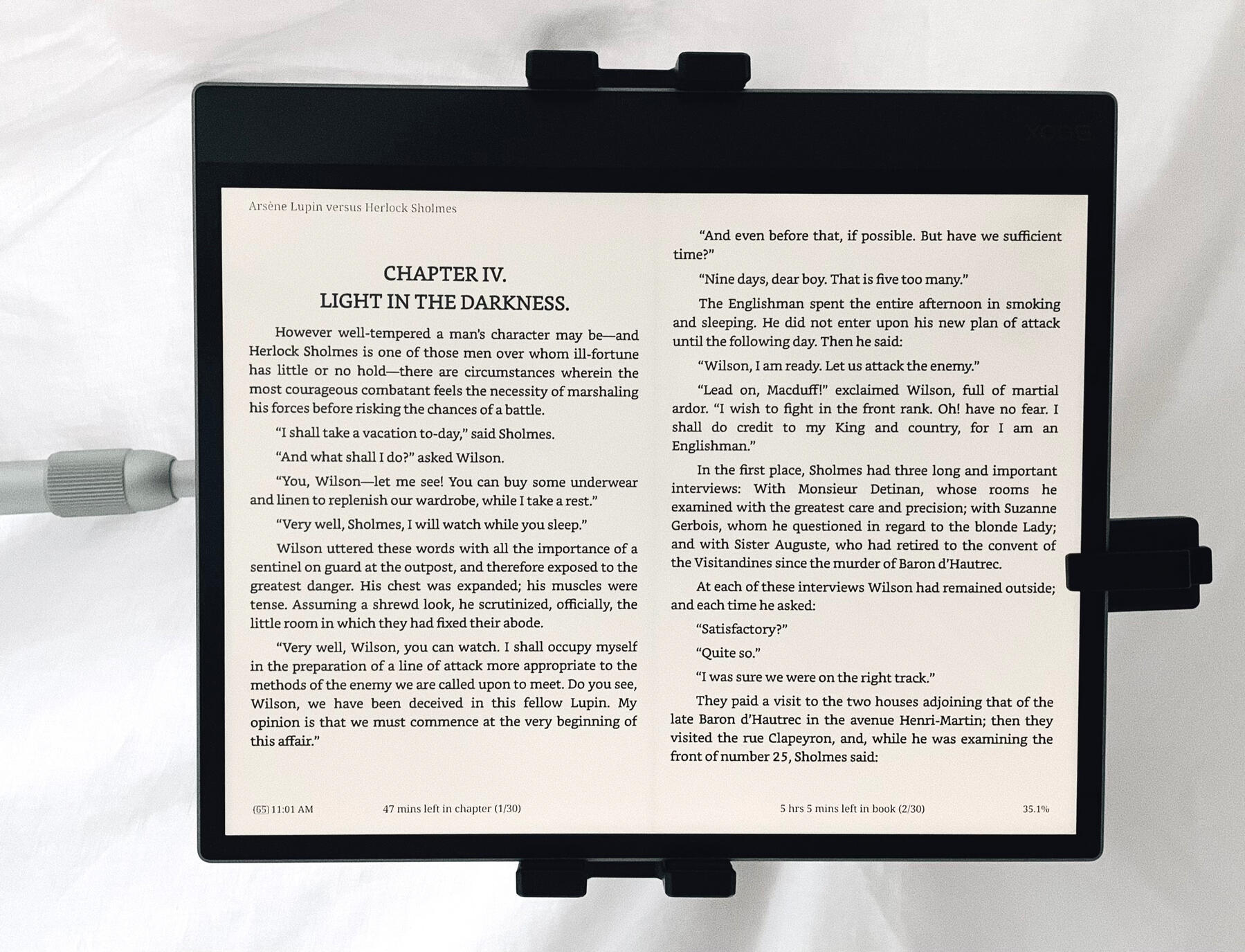 A large, black, e-ink Android tablet hangs in an arm-mounted clamp, with a remote-controlled page turner clipped to the right edge. The screen displays the first two pages of Chapter 4 of Arsène Lupin Versus Herlock Sholmes on a beige background.